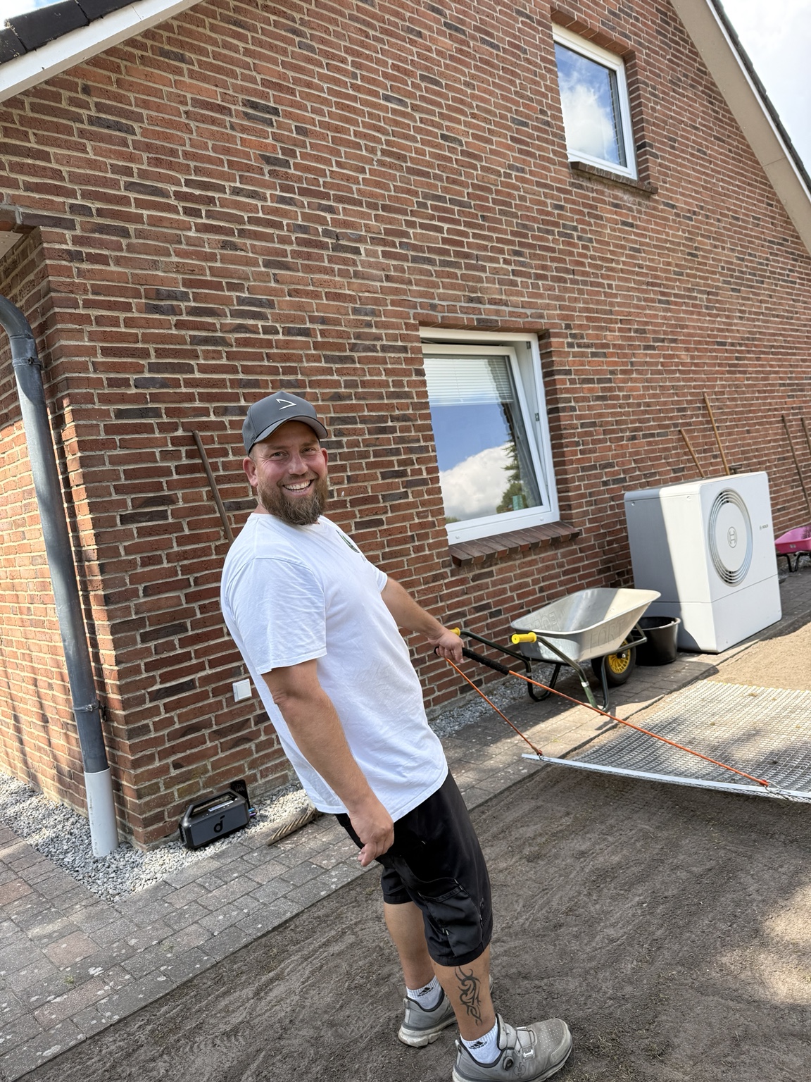 Matti mit Cap und weißem T-Shirt lächelt bei der Gartenarbeit in die Kamera